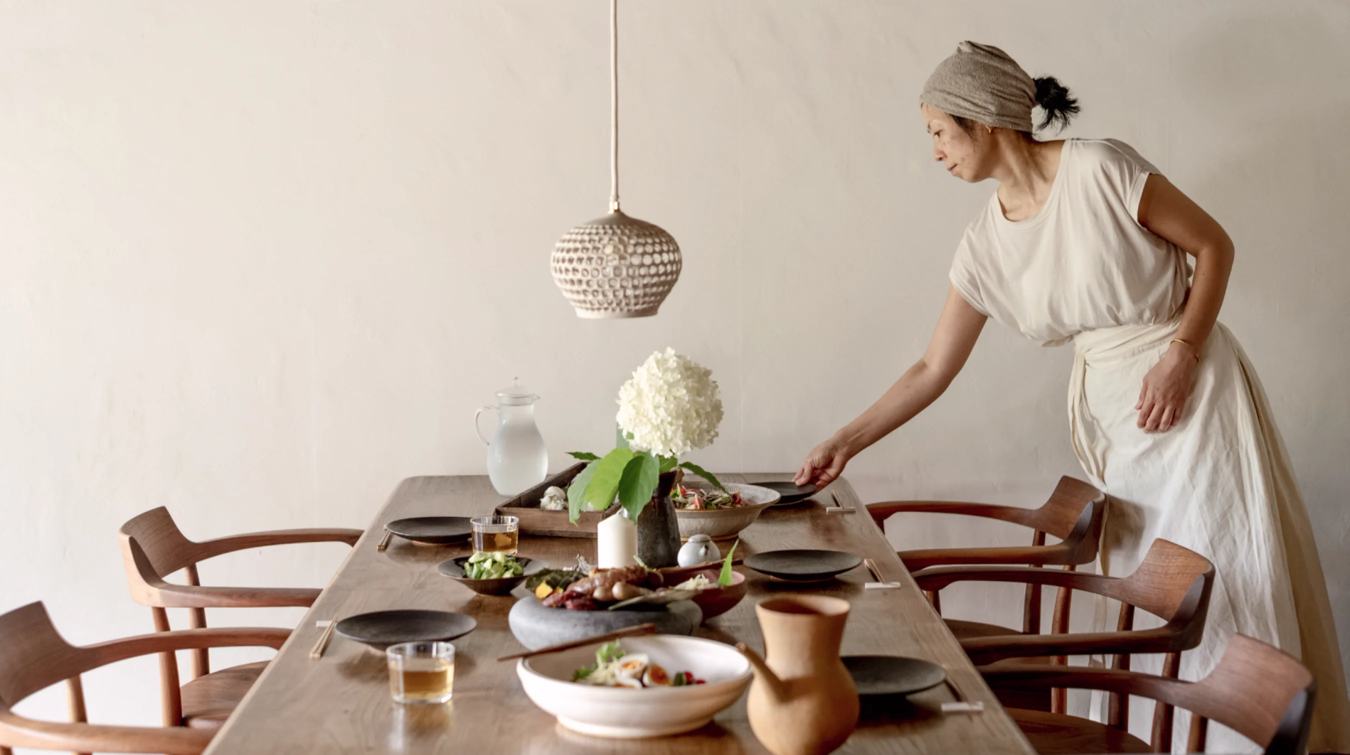 Minimalist dining room with long wooden table, woman setting dark plate, white hydrangea centerpiece