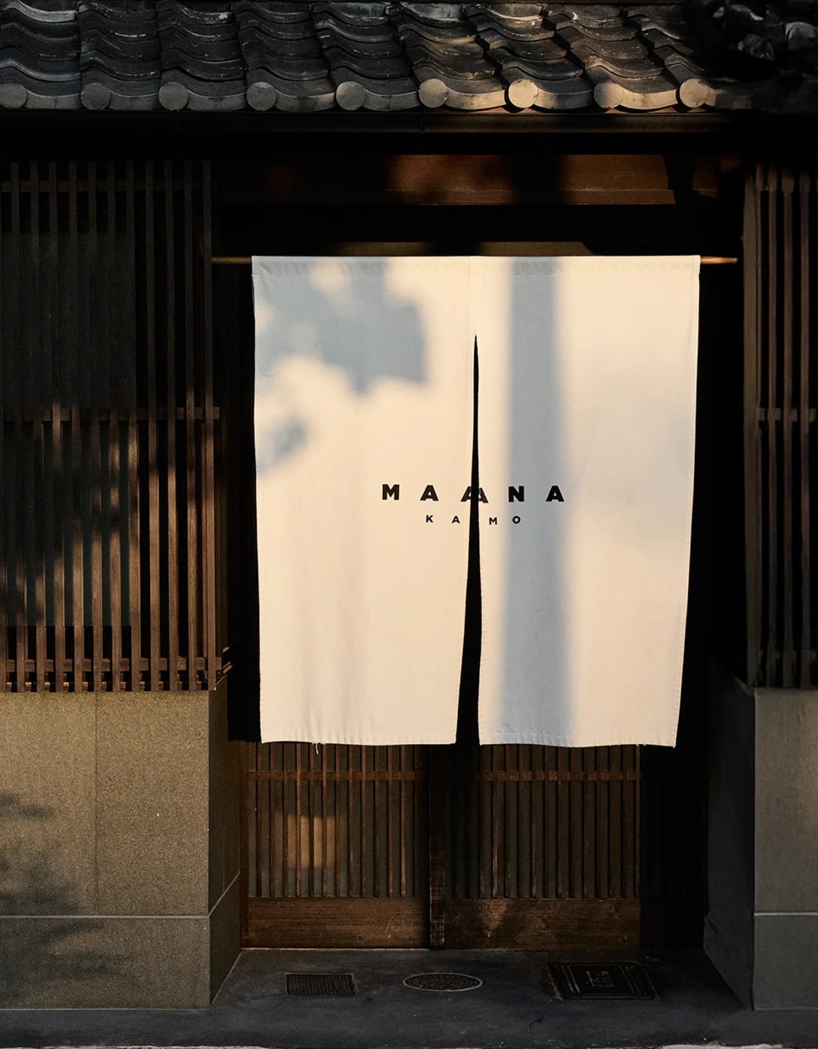 Maana Kamo exterior with wooden lattice facade, noren curtain, and stone lantern